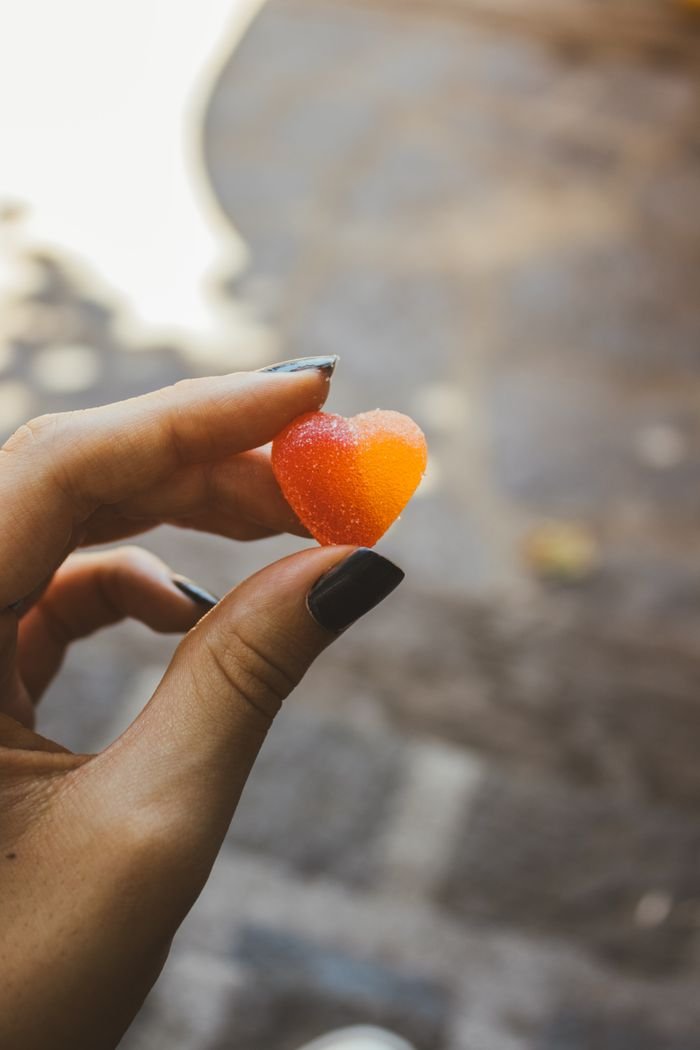 collection-01 A close-up of a hand holding a heart-shaped gummy candy with a blurred outdoor background.